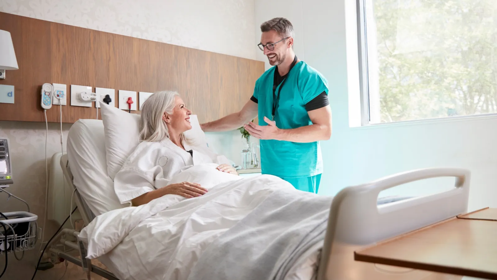 Male nurse talking to a smiling elderly woman patient in a bright hospital room by the window