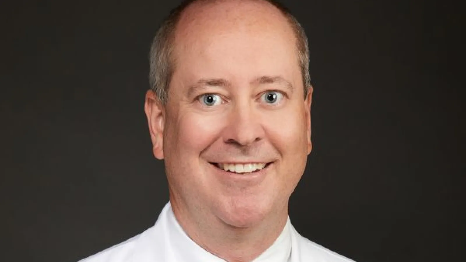 Smiling male orthopedic surgeon in white coat with Pinnacle Orthopaedics logo and gold tie on dark background