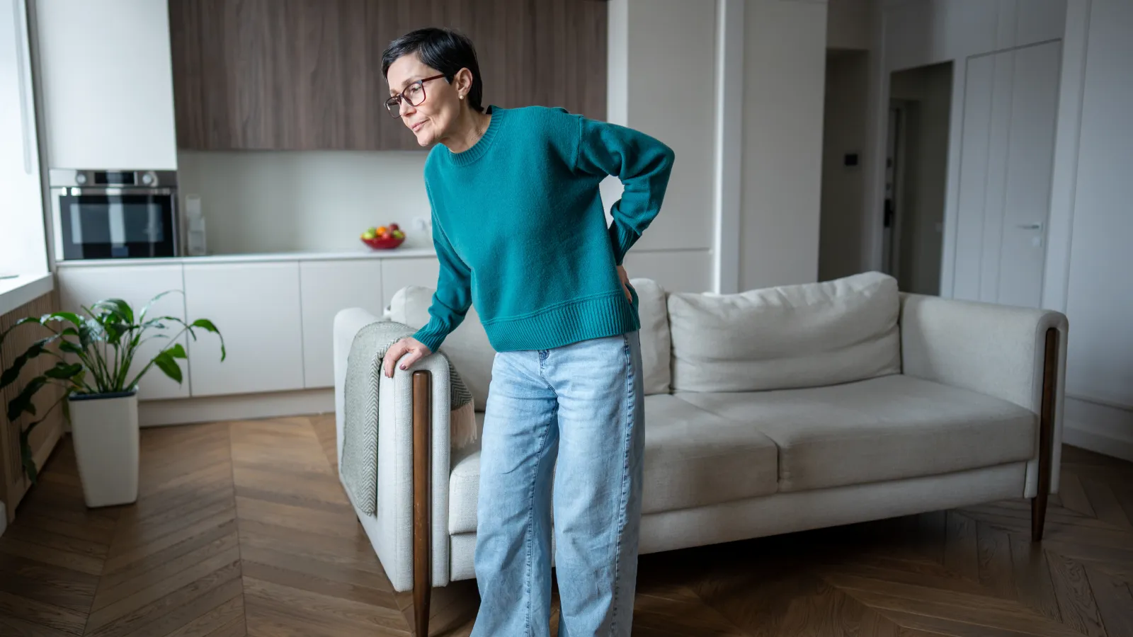 Woman in teal sweater and jeans holding her lower back, standing in a modern living room with a beige sofa.