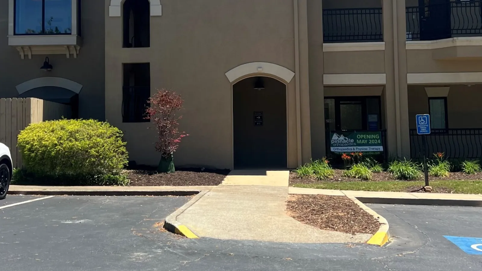 Two-story beige building with arched windows and entrances under a clear blue sky, surrounded by landscaping and parking.