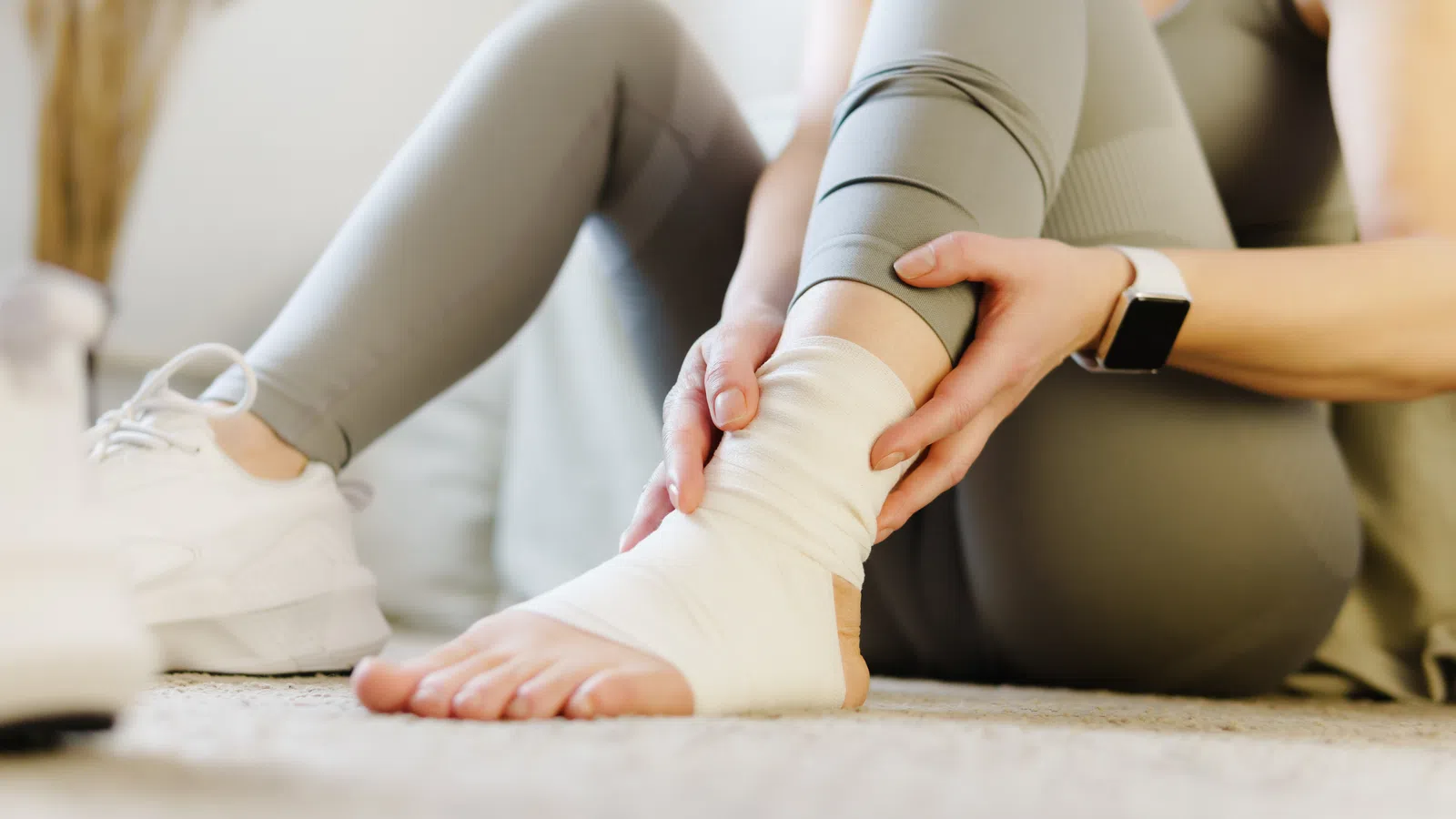 Person sitting on floor holding a wrapped ankle, wearing gray leggings and white sneakers indoors.