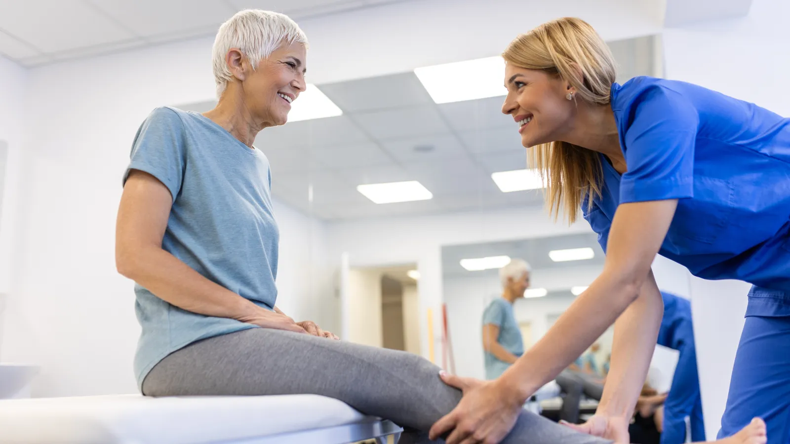 Older woman receiving physical therapy knee treatment from smiling therapist in modern clinic room.