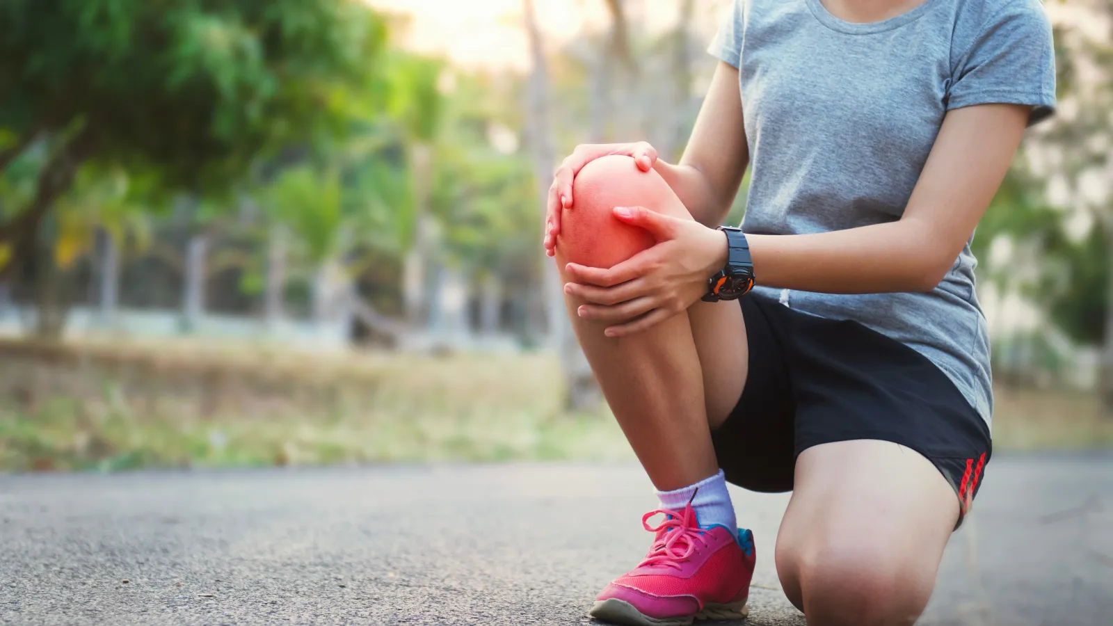 Person kneeling on pavement holding painful knee outdoors, wearing pink shoes and gray shirt during exercise.