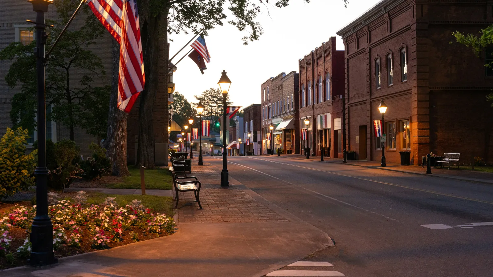 Evening view of a quiet small-town street with American flags, lampposts, benches, and historic brick buildings.