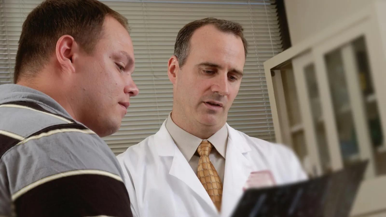 Doctor in white coat explaining medical results to a patient in a consultation room with blinds in background