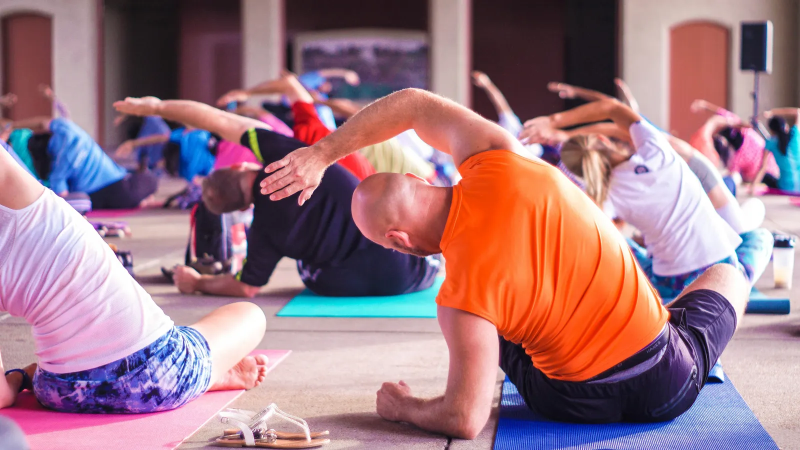 Group of people performing side stretches on yoga mats in a spacious indoor class with vibrant athletic wear.