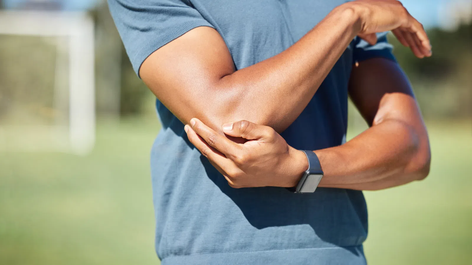 Man in blue shirt holding his elbow outdoors, indicating elbow pain or injury.