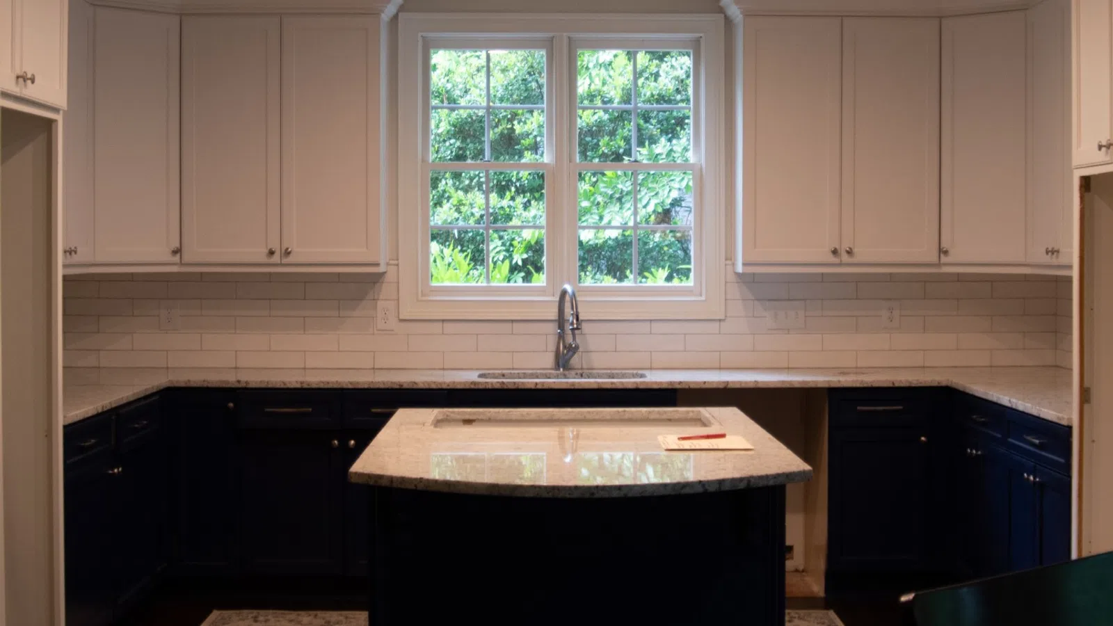 Modern kitchen with white upper cabinets, navy blue lower cabinets, center island, and large window with greenery view