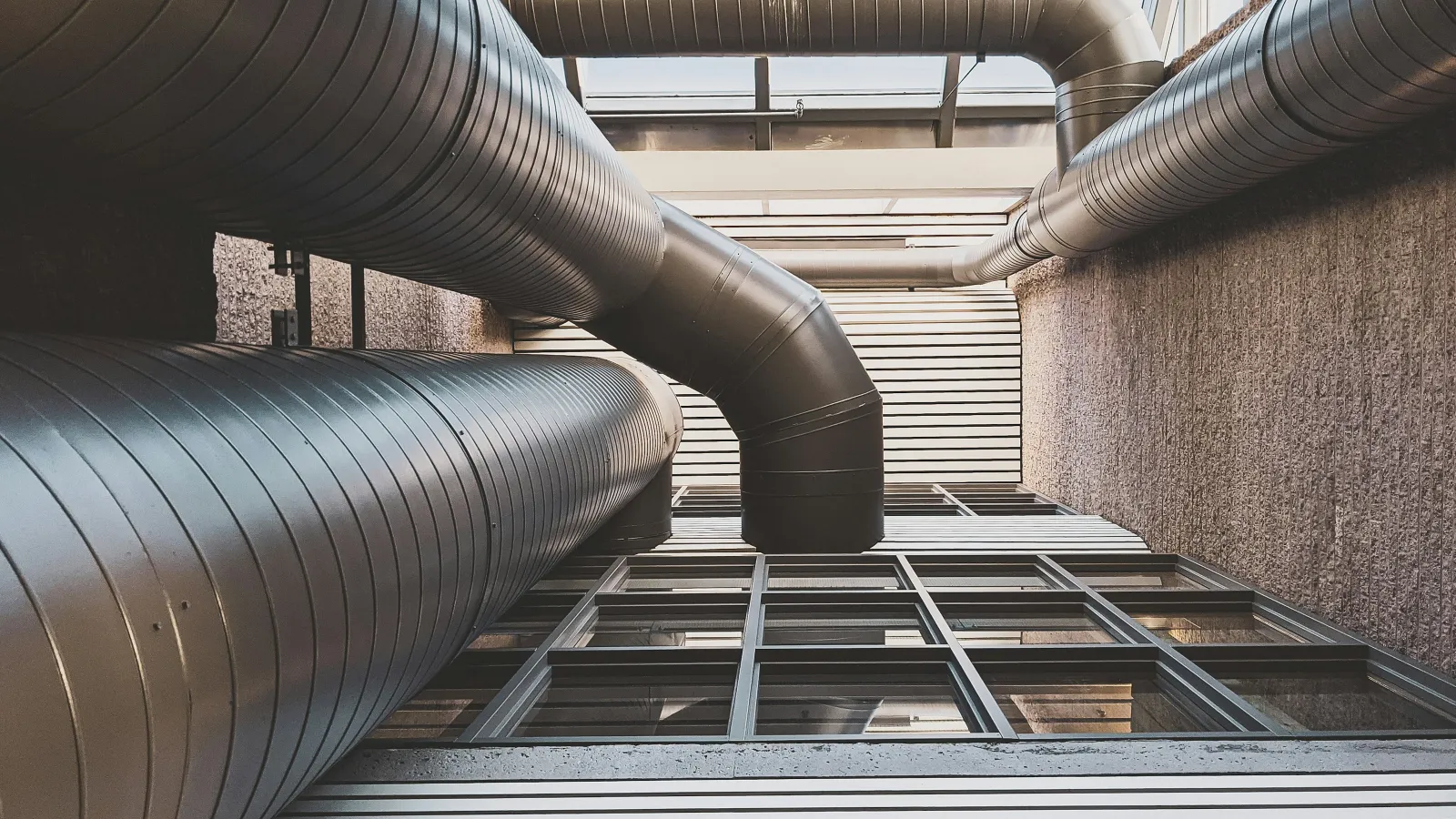 Upward view of large industrial ventilation ducts attached to a building with windows and textured walls.