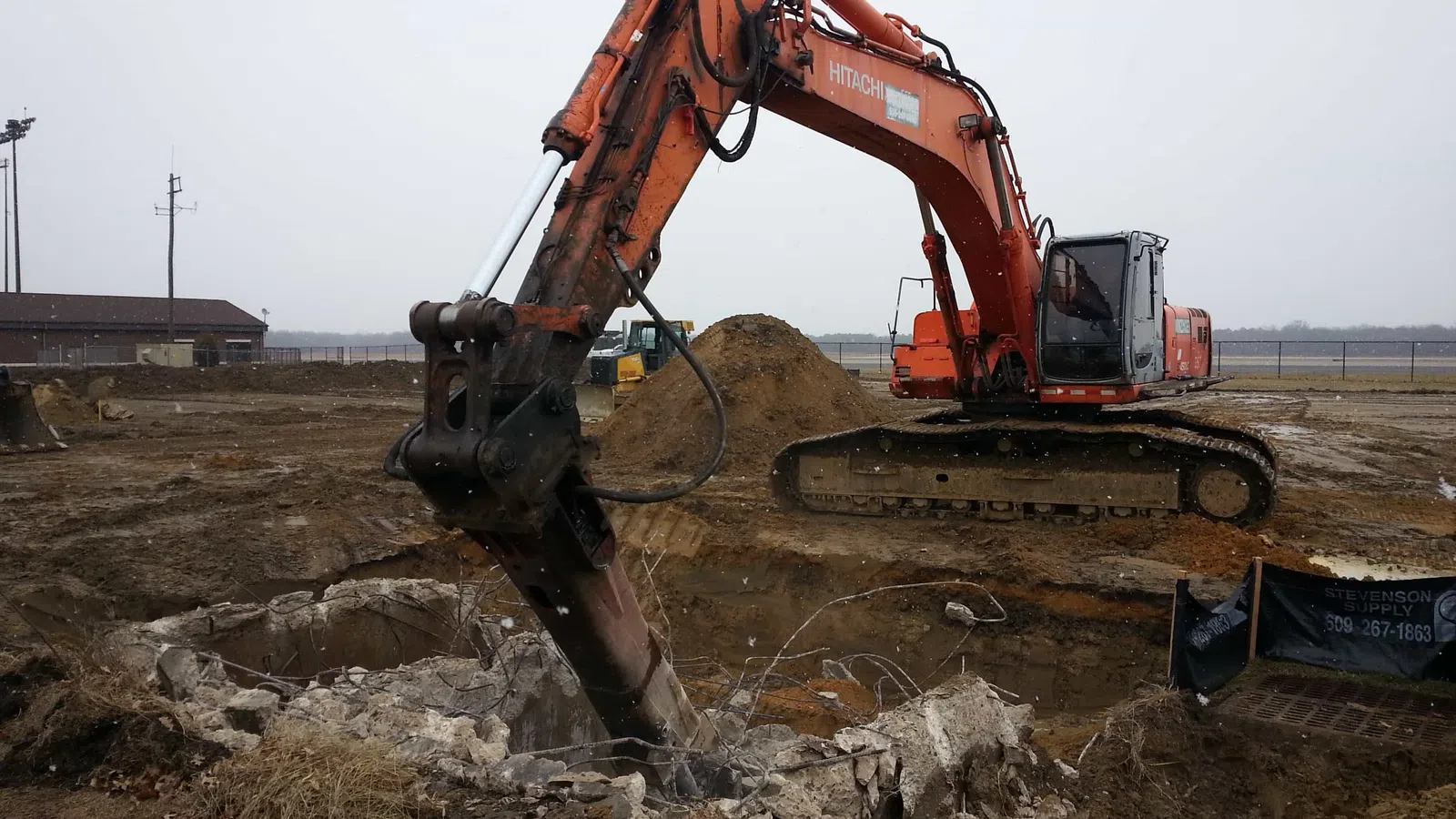 Orange excavator breaking concrete at a muddy construction site under overcast sky.