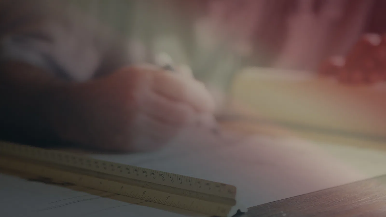 Person sketching architectural plans with a ruler and pencil on a cluttered desk in soft light.
