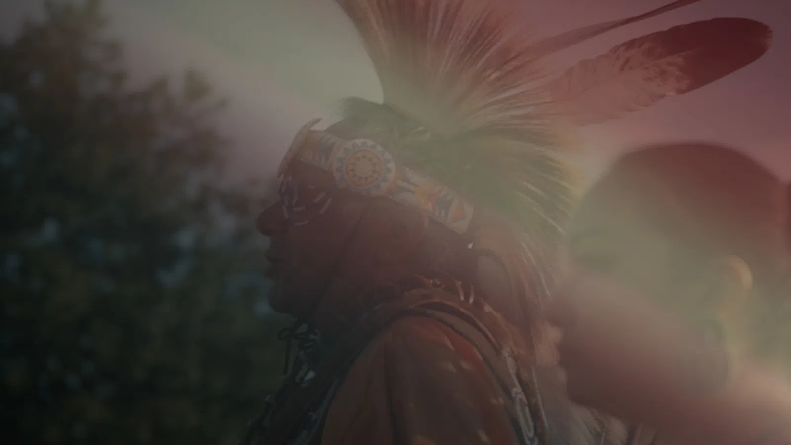 Silhouetted Native American man with feathered headdress at sunset with soft sunlight and blurred background.