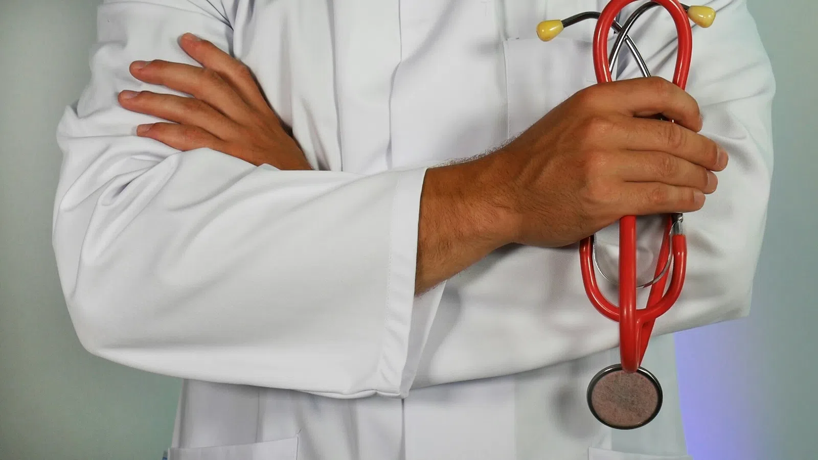Doctor wearing white coat with arms crossed holding a red stethoscope against light background