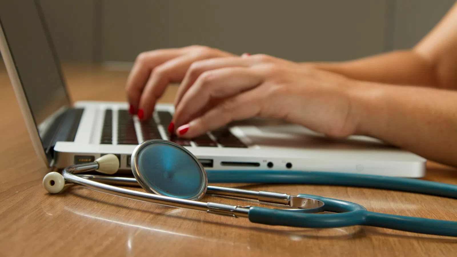 Hands typing on a laptop keyboard next to a stethoscope on a wooden table symbolizing medical work.