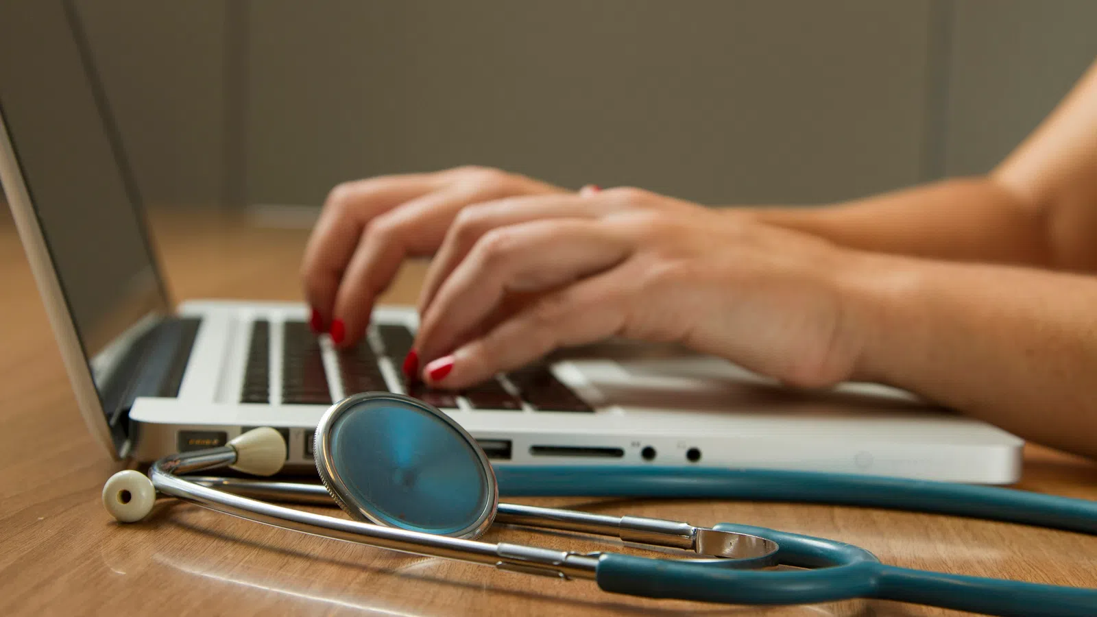 Hands typing on a laptop keyboard with a stethoscope resting on the wooden table nearby.