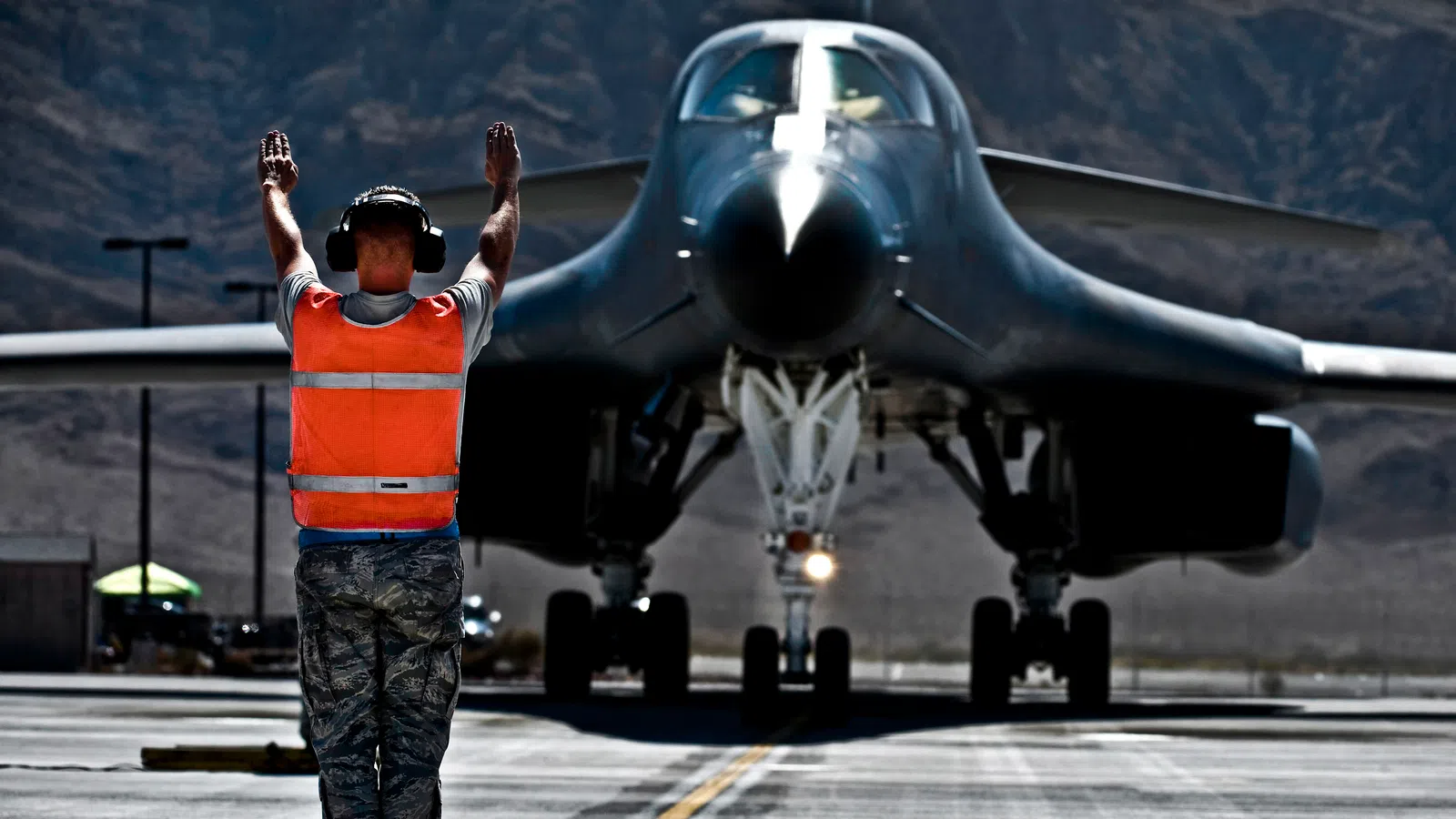 Ground crew member directing a large military aircraft on a runway in a mountainous desert area.
