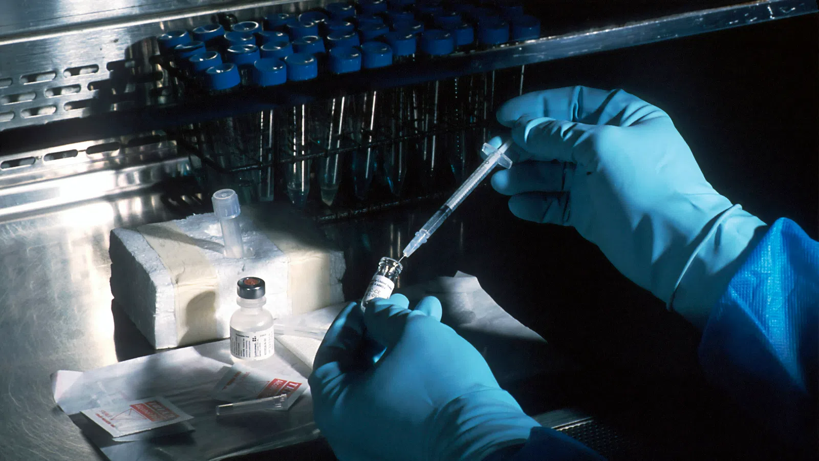 Scientist in blue gloves prepares vaccine vial with syringe inside a sterile lab environment.