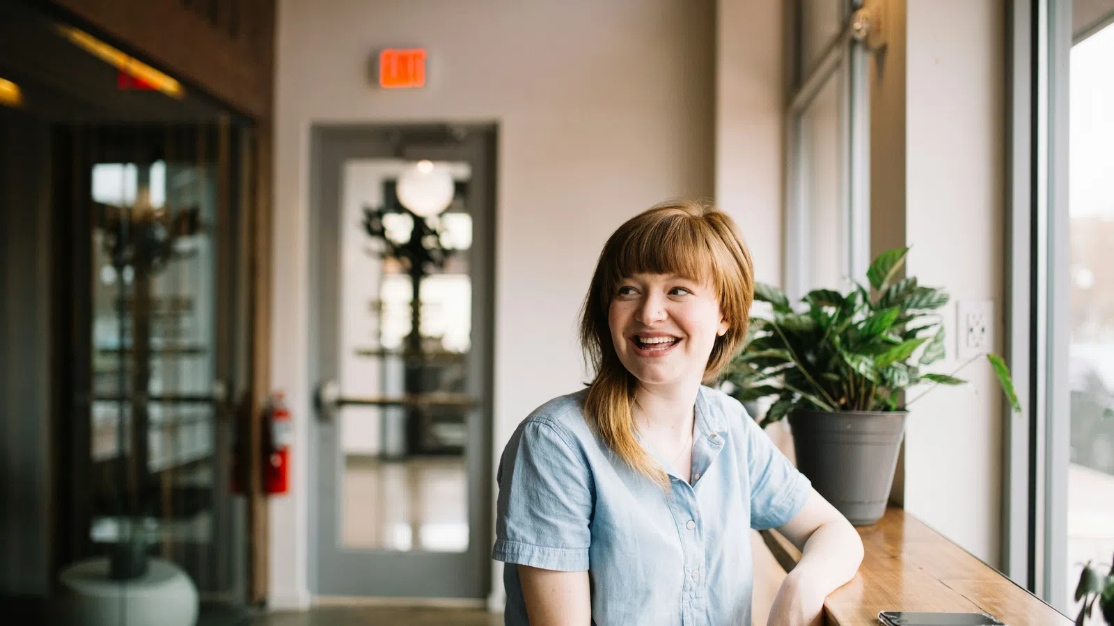 Smiling young woman in blue shirt sitting by a window in a bright modern cafe with plants and phone on table
