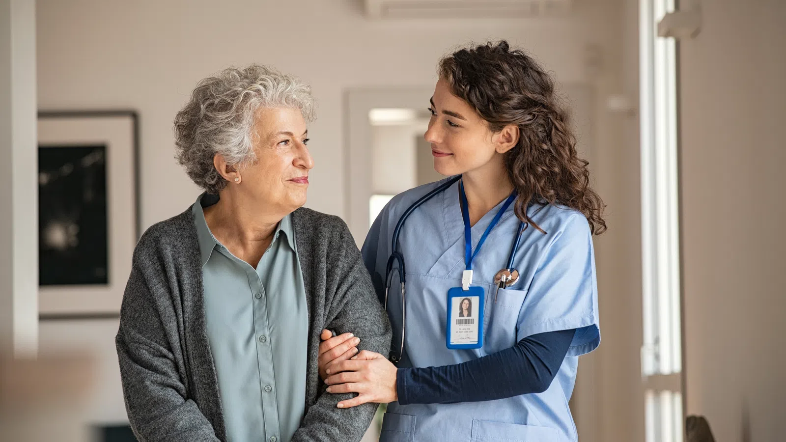 Nurse gently supporting elderly woman as they walk indoors, showing care and compassion in a home setting.