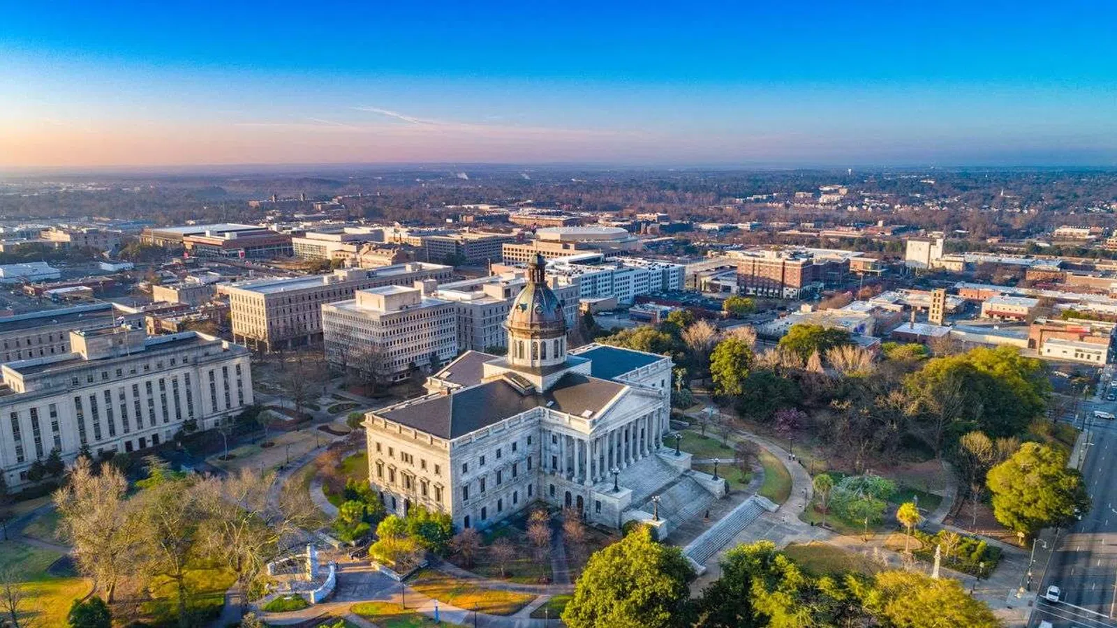 Aerial view of a historic capitol building surrounded by greenery and cityscape under a clear blue sky.
