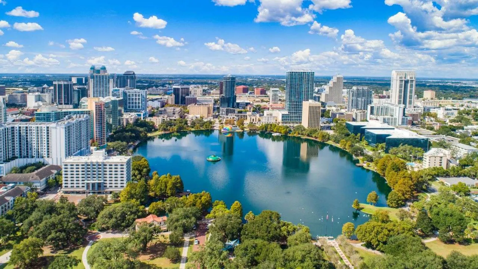 Aerial view of downtown Orlando with Lake Eola surrounded by high-rise buildings and green parks under a blue sky.