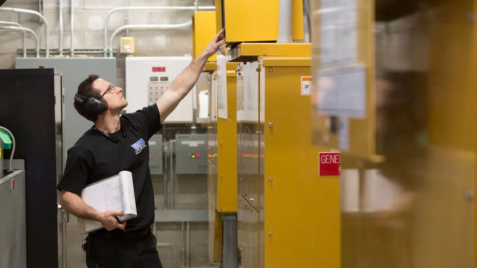 Technician wearing headphones inspects yellow industrial control panel while holding documents in a utility room.