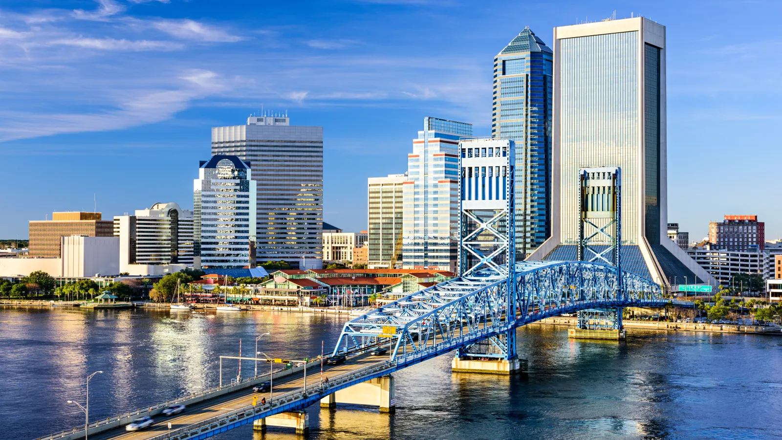 Blue steel bridge over river with city skyline and tall buildings under a clear blue sky