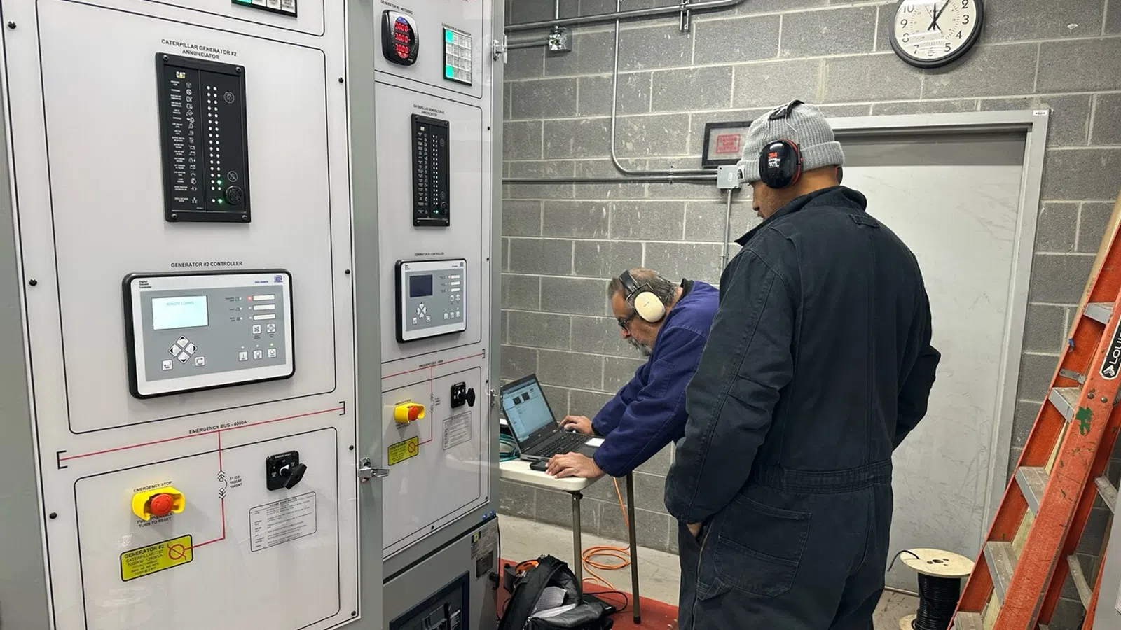 Two technicians wearing headphones inspecting electrical control panels and working on a laptop in an industrial room.