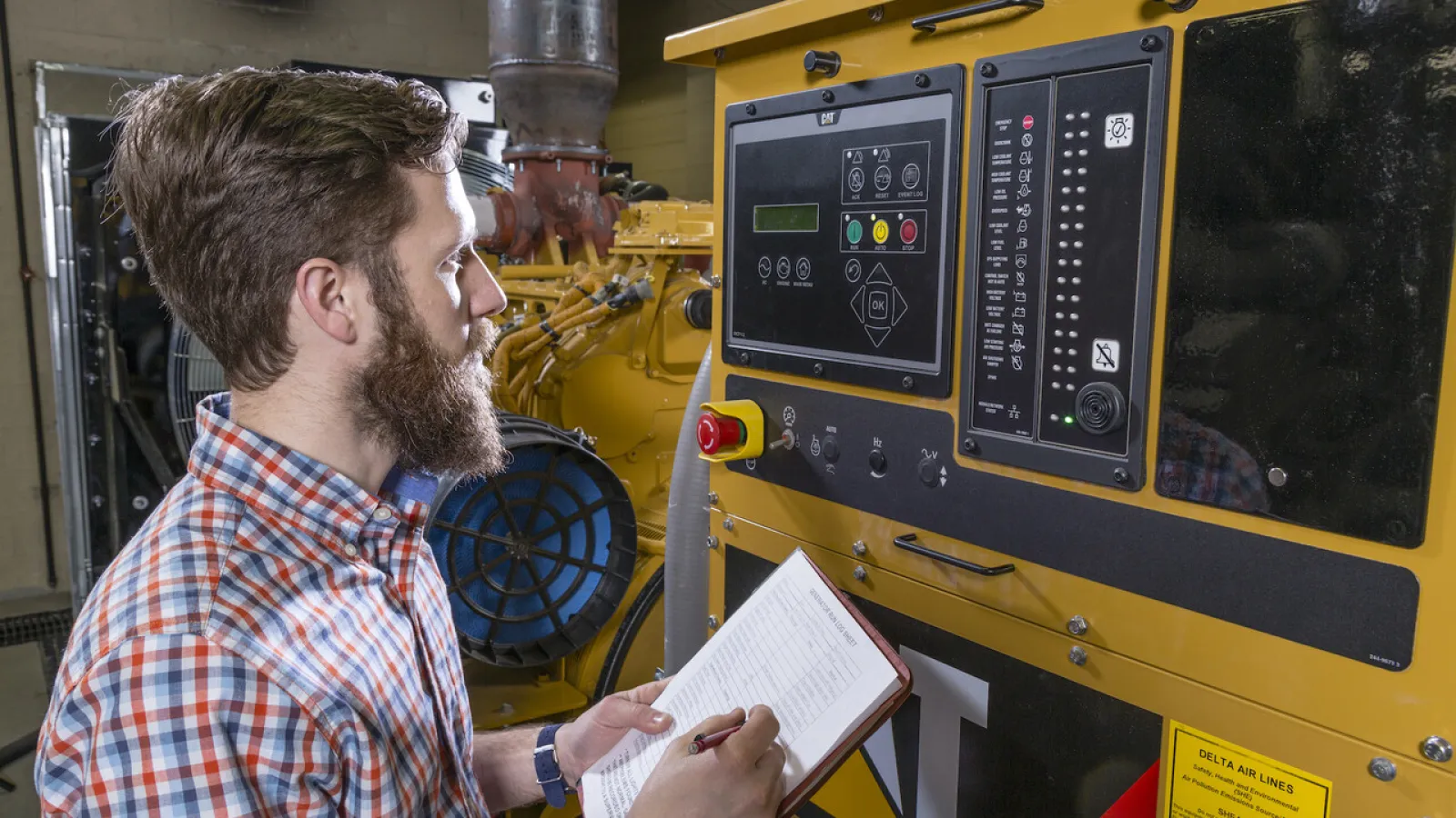 Man in plaid shirt monitoring and taking notes on large industrial CAT machine control panel in factory.
