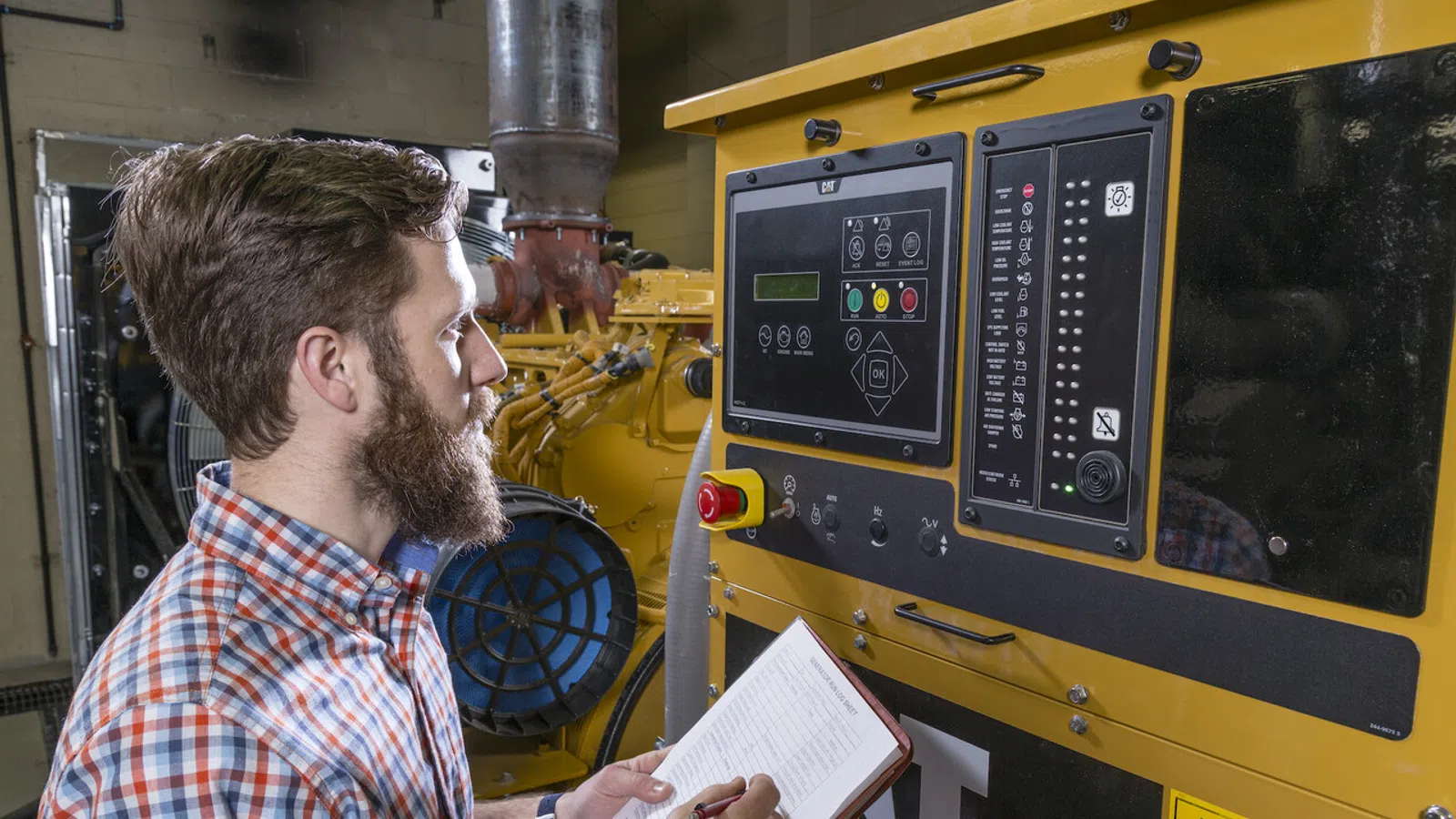 Man in plaid shirt monitoring and taking notes on large industrial CAT machine control panel in factory.