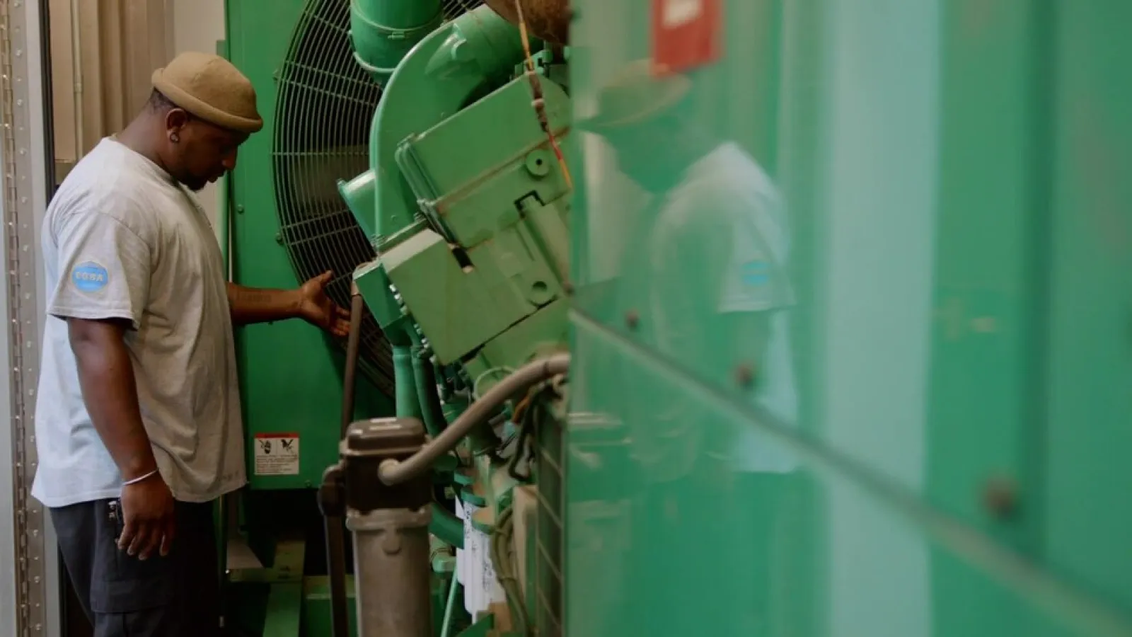 Technician inspecting large green industrial machinery in a mechanical room with safety signage visible