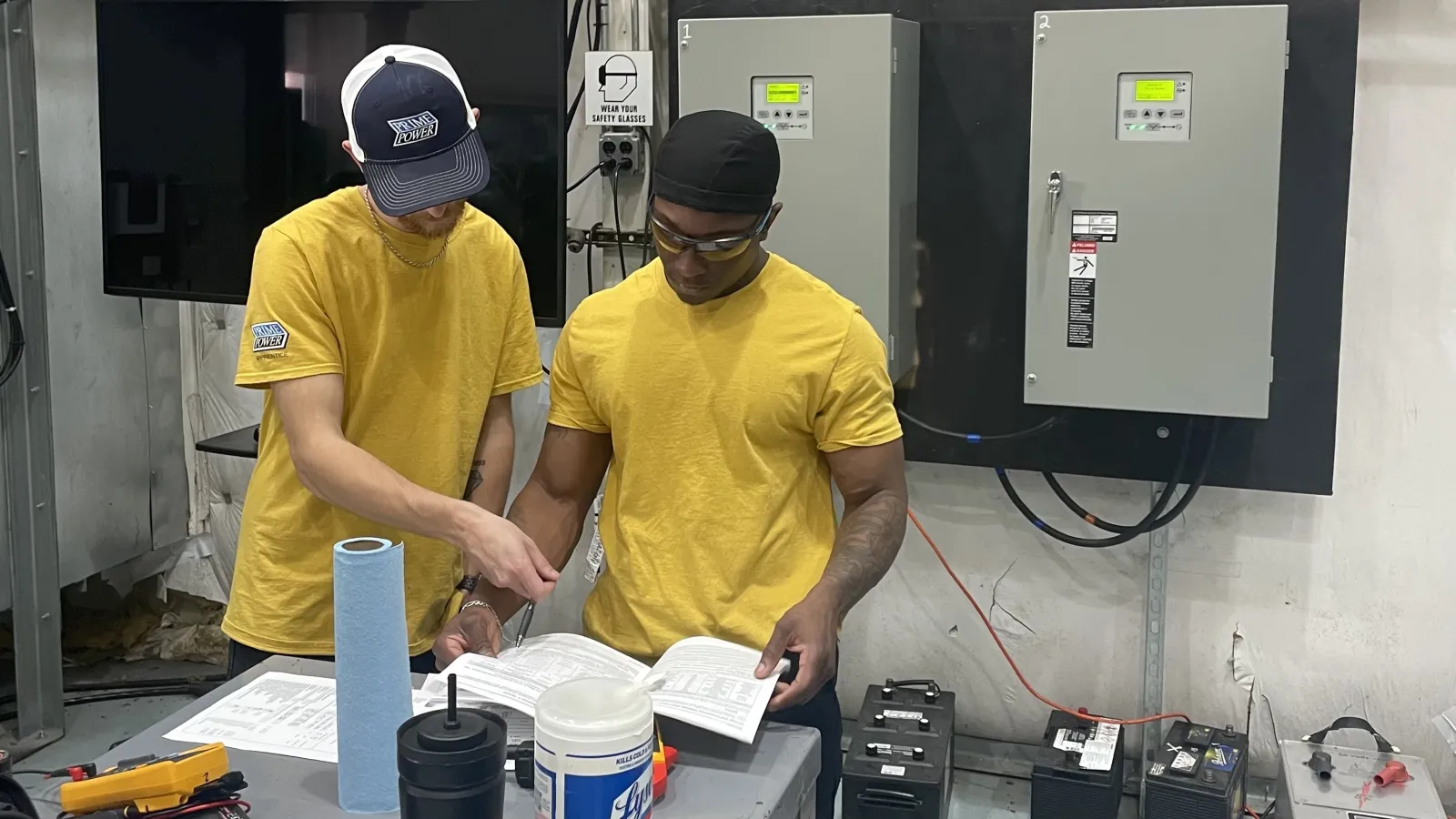Two men in yellow shirts reviewing paperwork at a workbench in an industrial setting with electrical panels.