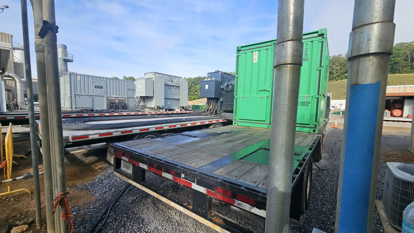 Flatbed trailer with wooden planks and a green shipping container in an industrial outdoor area on a sunny day
