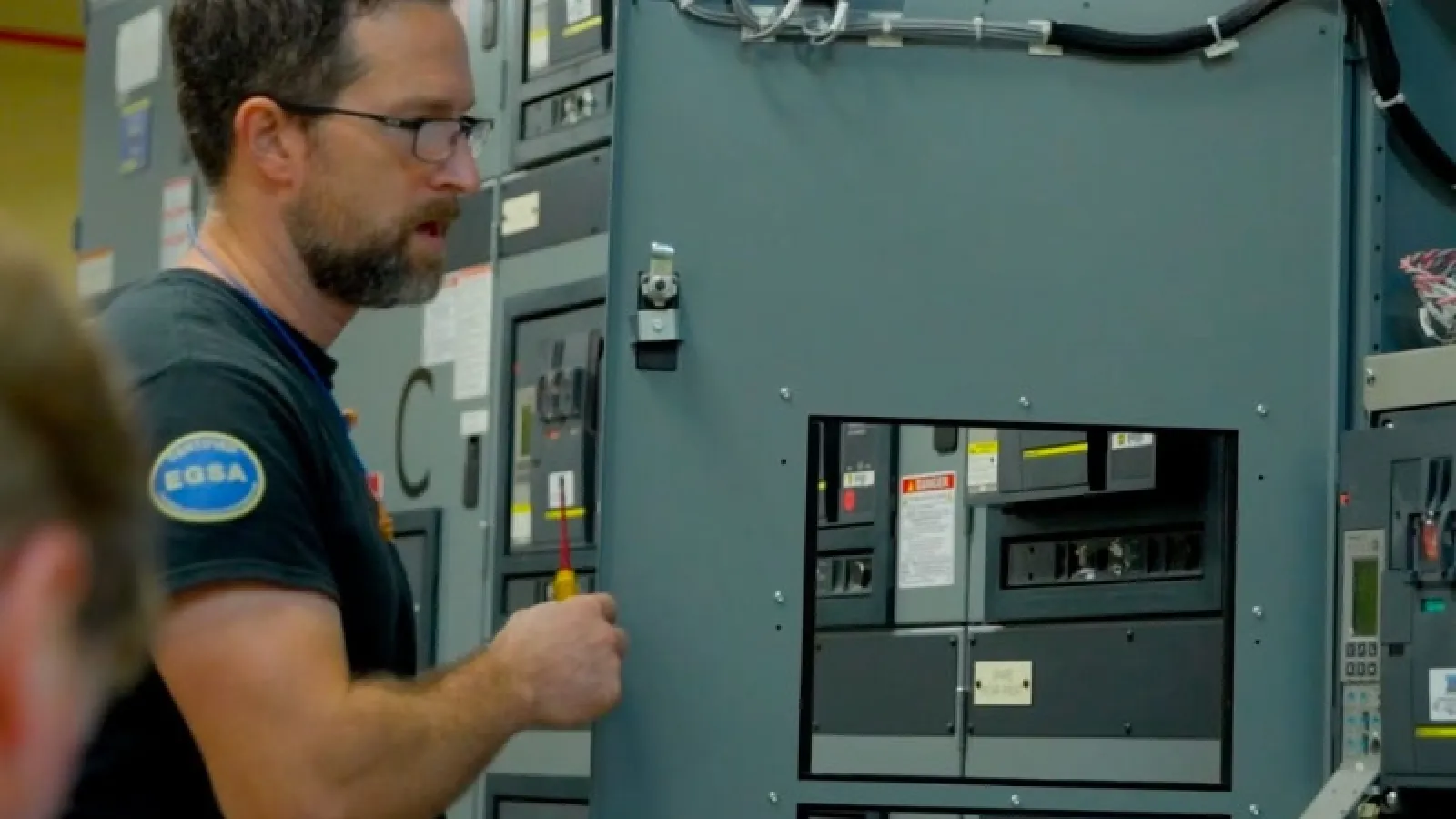 Technician wearing glasses inspecting and working on electrical control panels in an industrial setting
