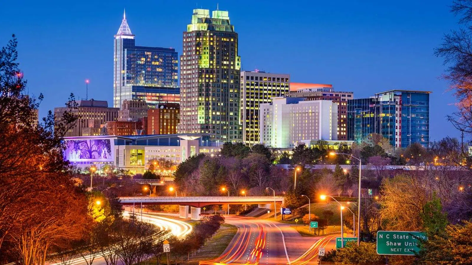 Nighttime cityscape of Raleigh, North Carolina with illuminated downtown buildings and light trails on the highway.