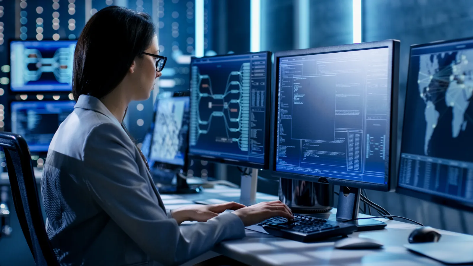 Woman in lab coat working on multiple computer screens with data and network diagrams in a technology lab.