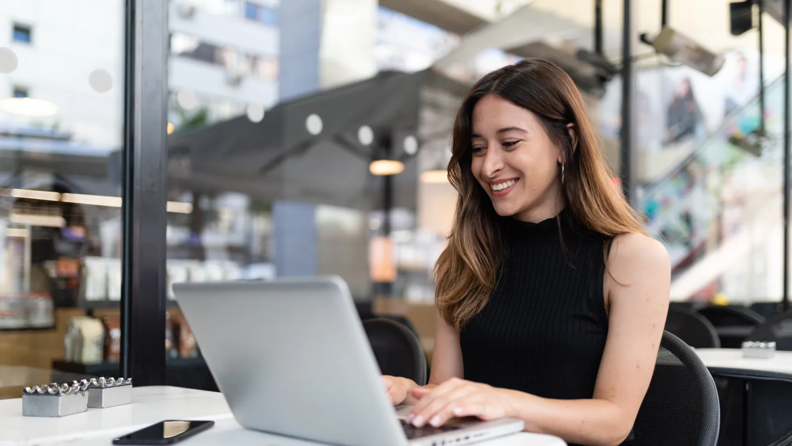 Smiling woman working on laptop at a modern cafe table with smartphone nearby in a bright atmosphere
