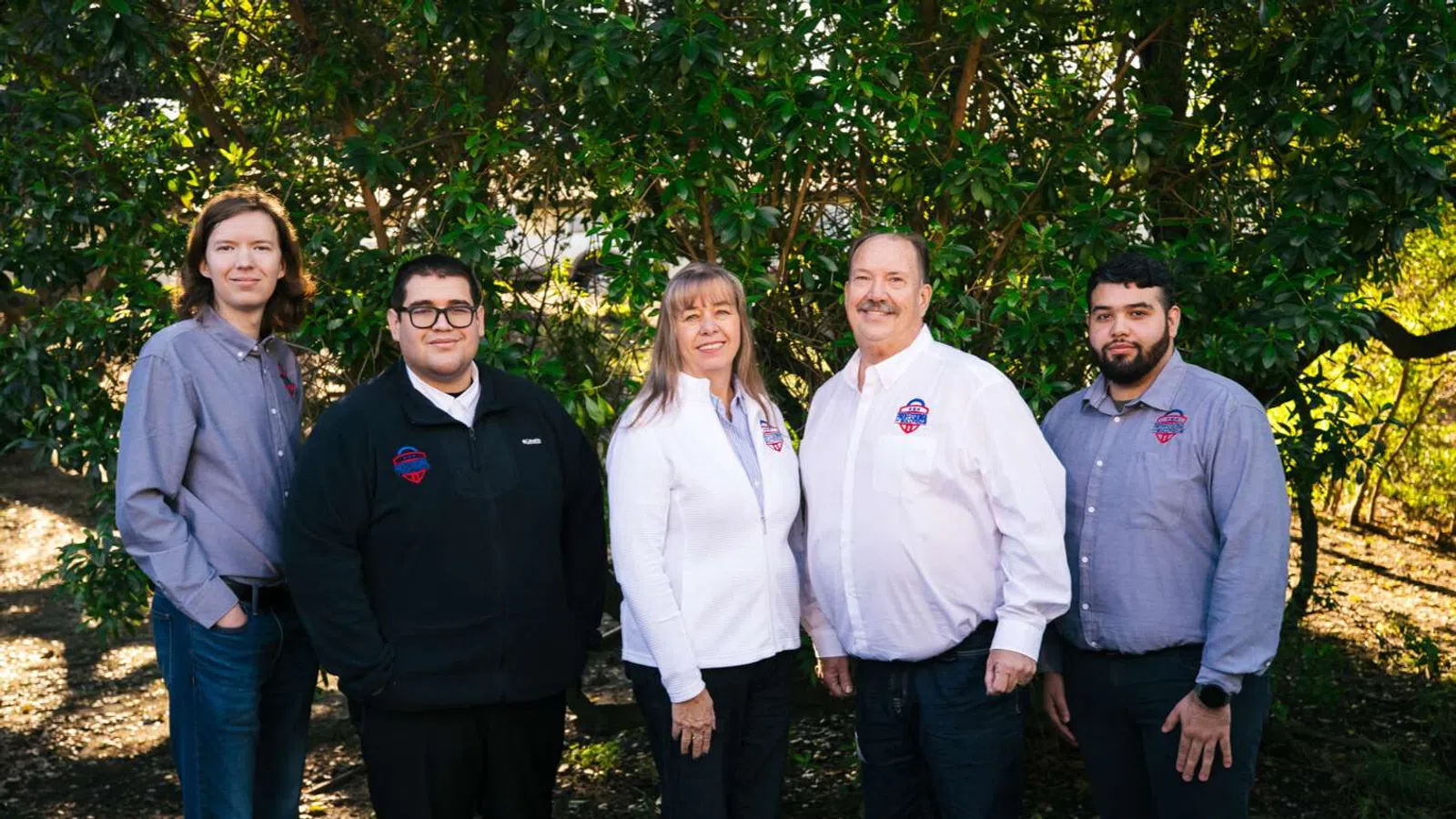 Group of five professionals standing outdoors under trees, wearing business casual and company-branded shirts.