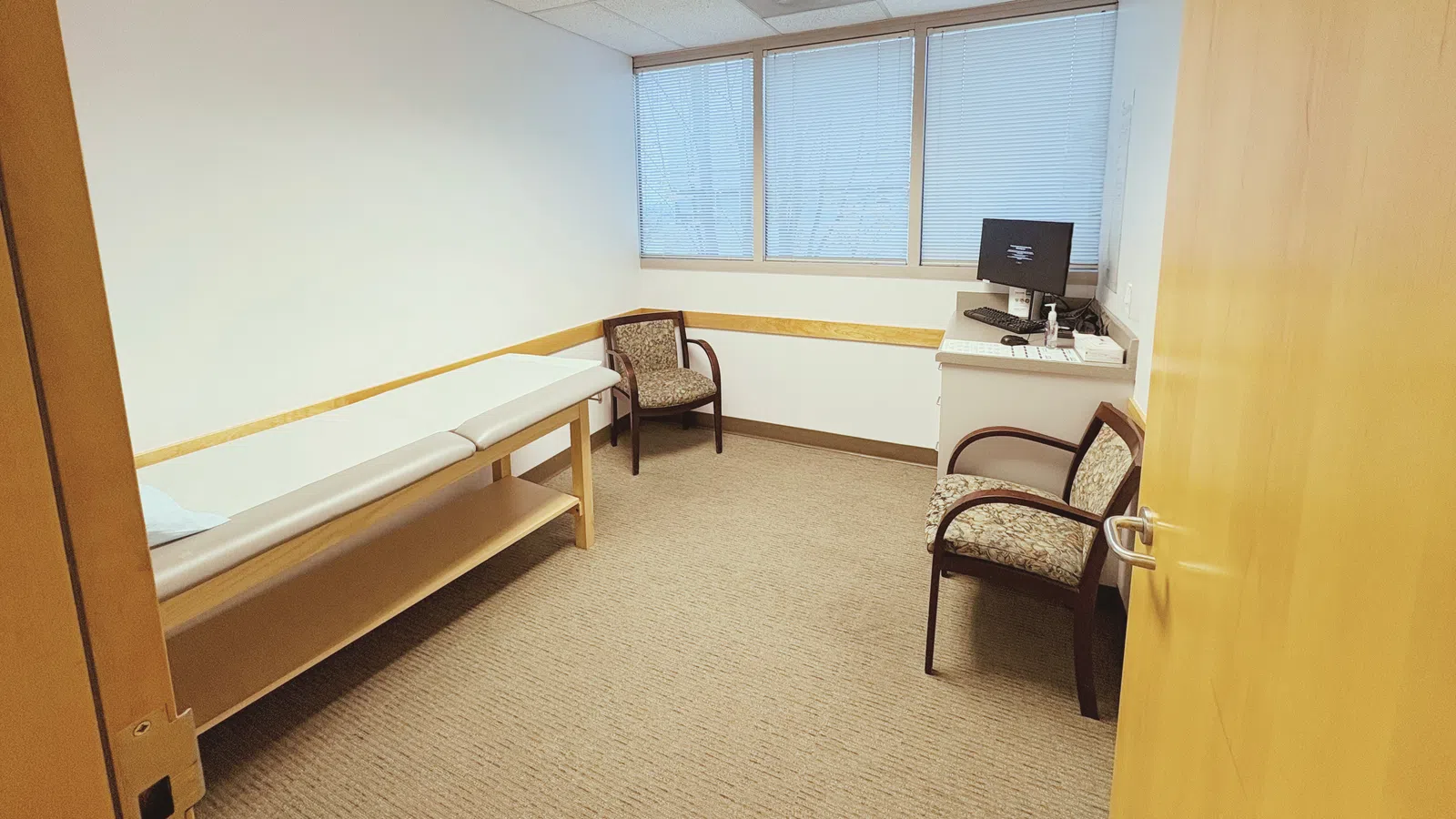 Bright medical examination room with two chairs, exam table, computer desk, and overhead fluorescent light.