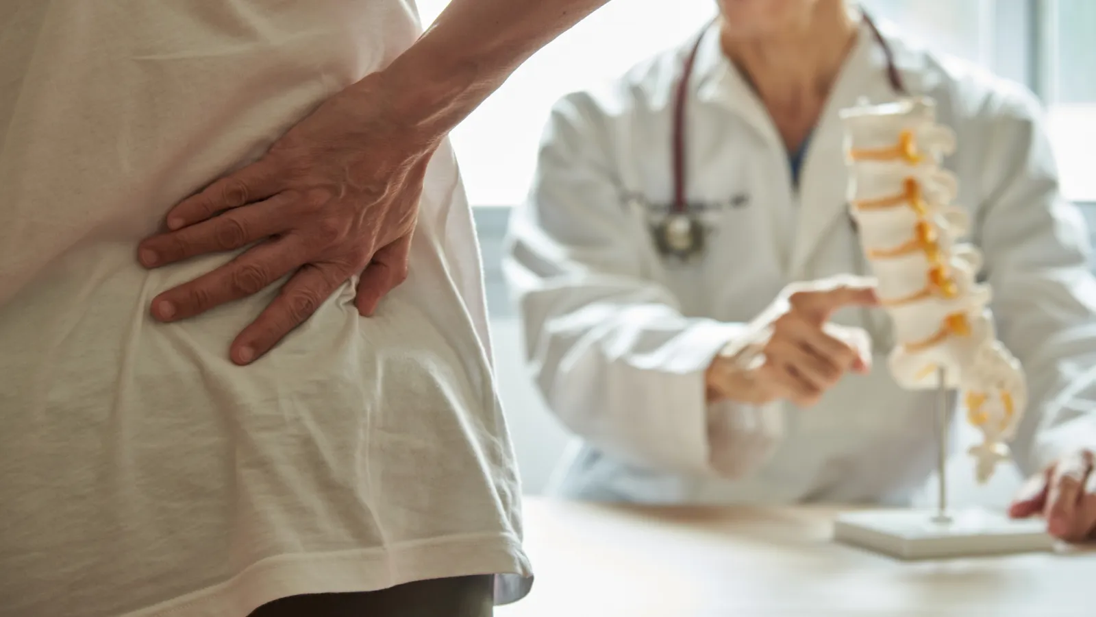 Patient holding lower back pain while doctor discusses spinal health using a spine model