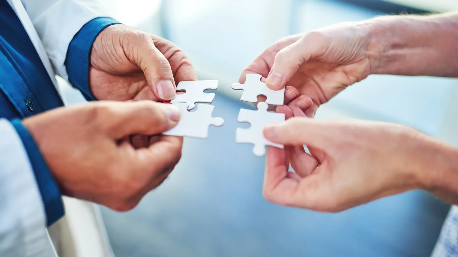 Four hands holding white puzzle pieces about to connect symbolizing teamwork and collaboration