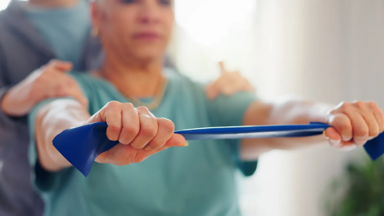 Senior woman doing resistance band exercise with physical therapist support indoors to improve strength.
