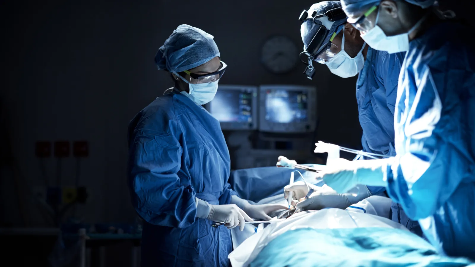 Surgeons in blue scrubs perform an operation in a dimly lit operating room with medical monitors in the background