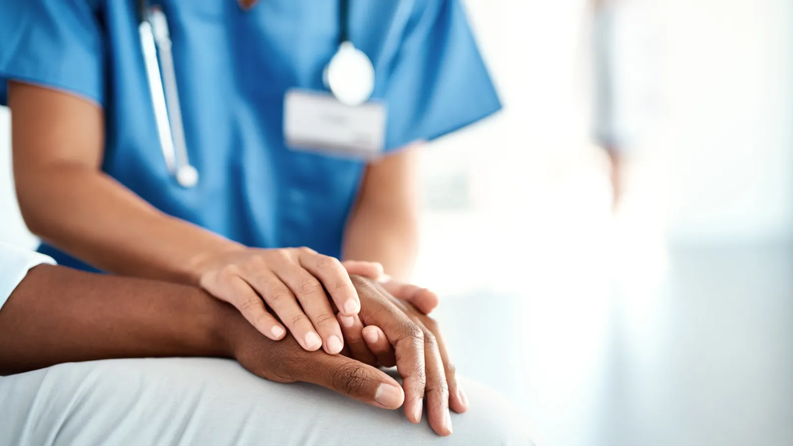Healthcare professional in blue scrubs comforting patient by holding their hand in a medical setting.