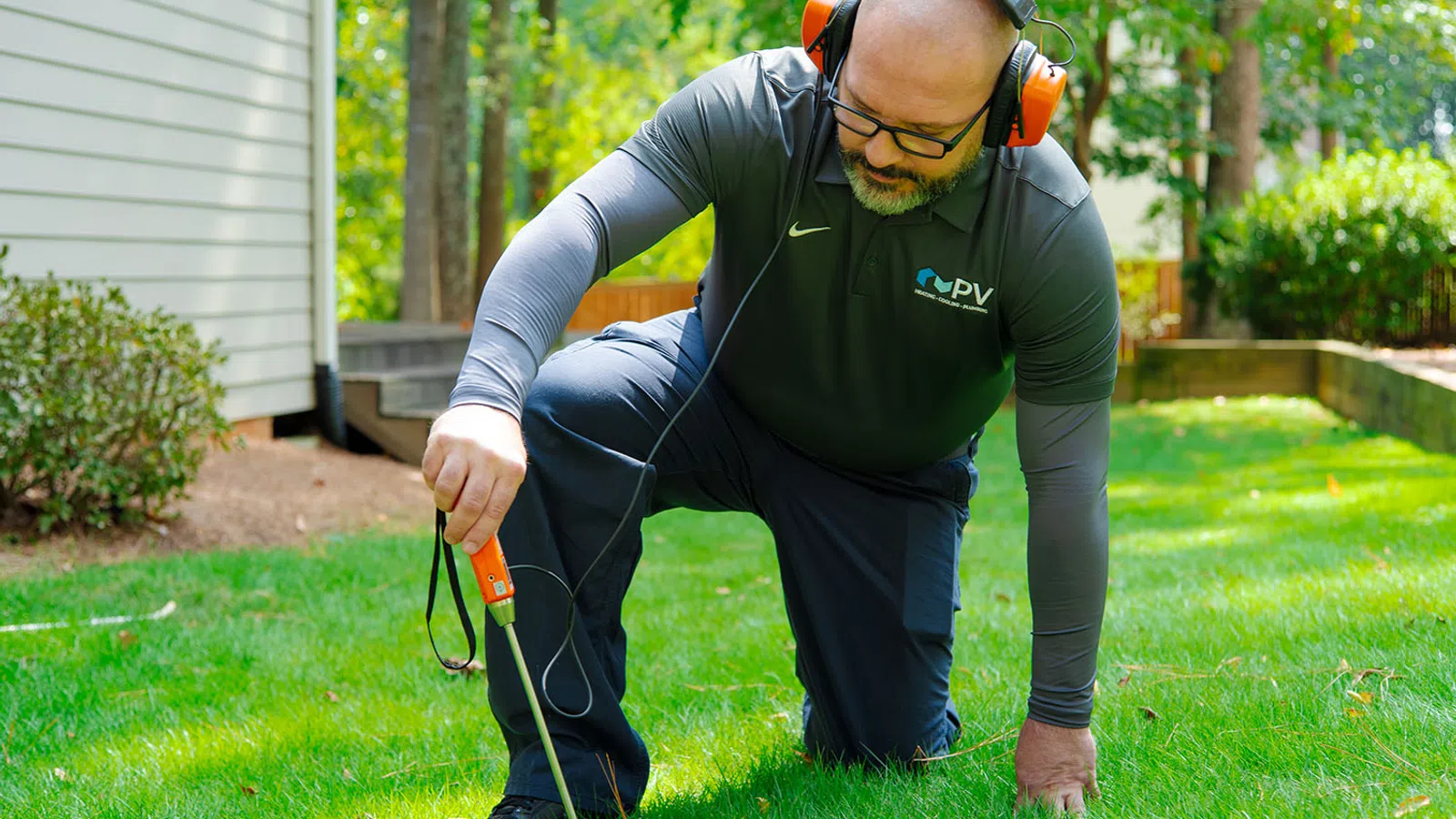 Man wearing ear protection and glasses kneeling on grass using a handheld device for soil or cable detection near a house.
