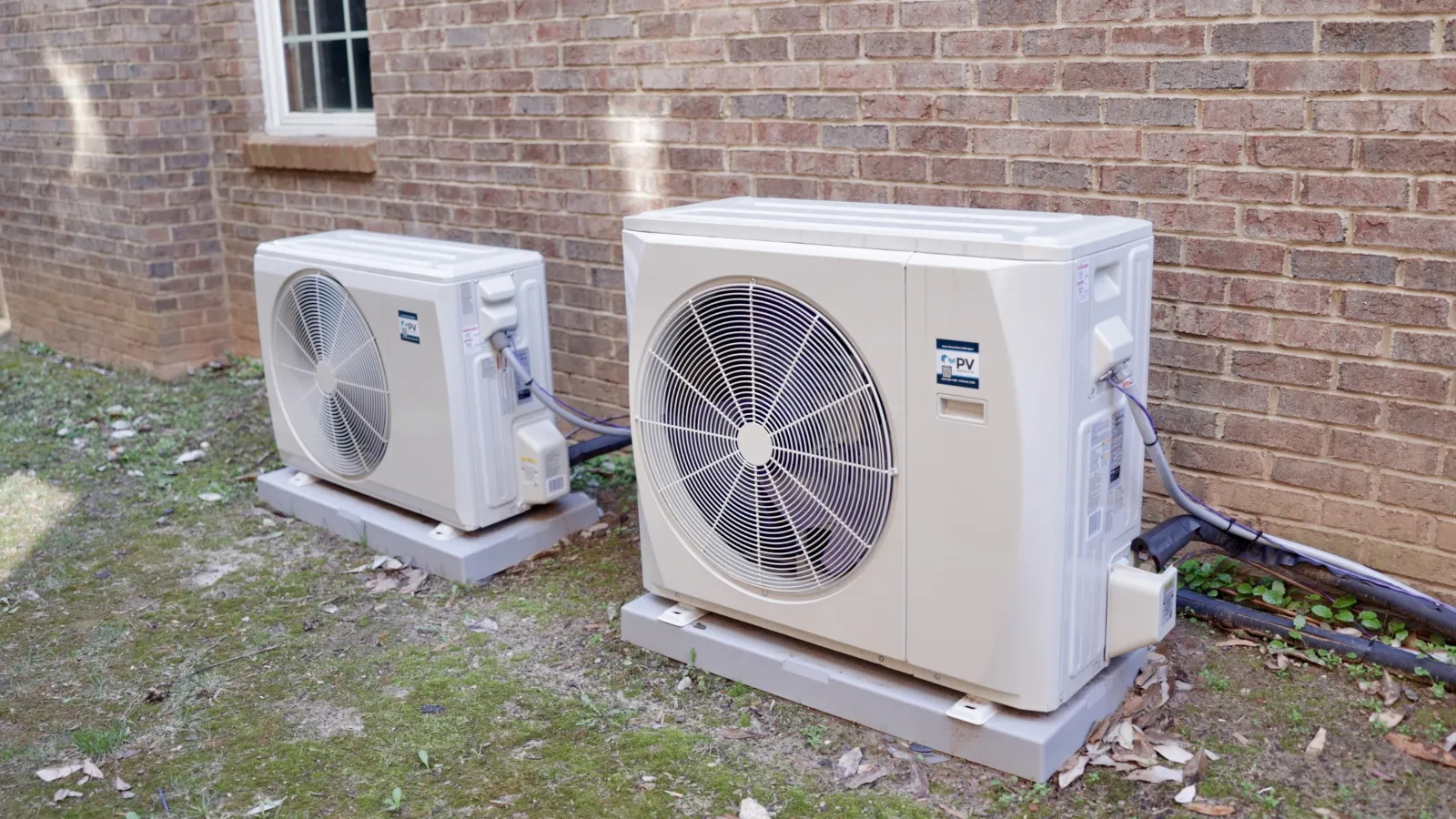Two outdoor mini-split air conditioning units installed on concrete pads against a brick house wall with grassy ground.