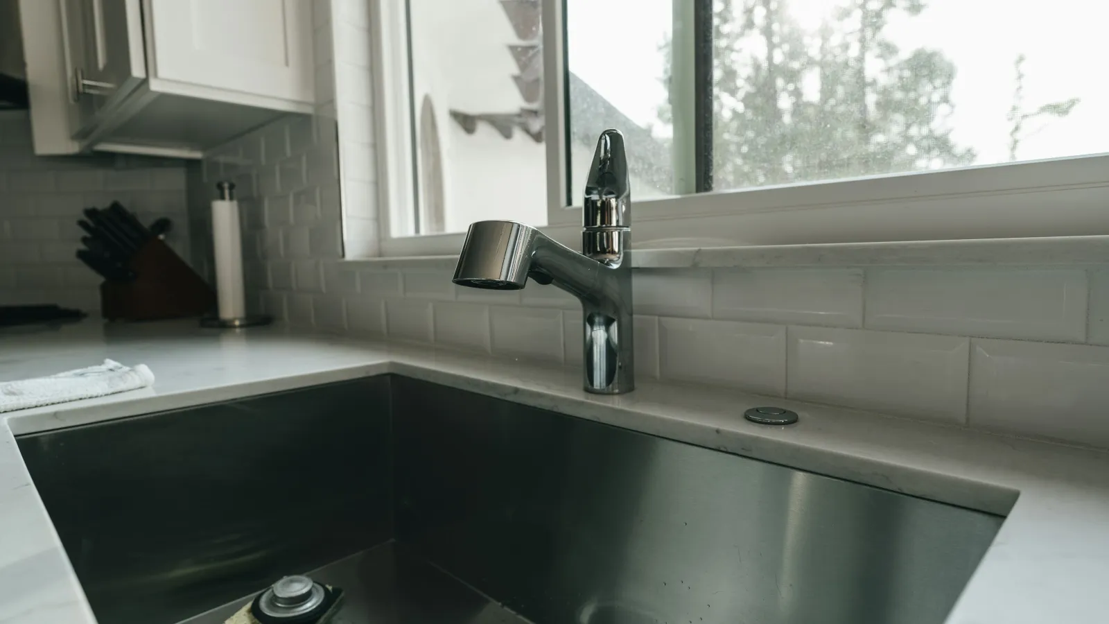 Modern kitchen sink with stainless steel basin and chrome faucet under a bright window with white tile backsplash.