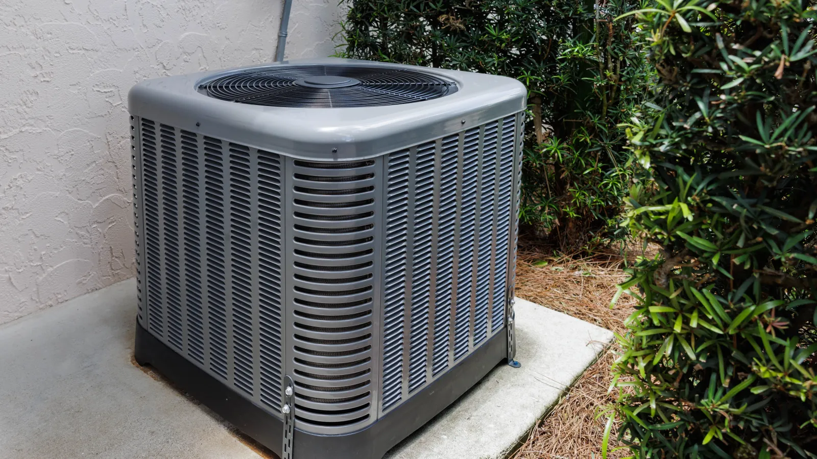 Outdoor gray air conditioning unit installed on concrete pad next to house and bushes.