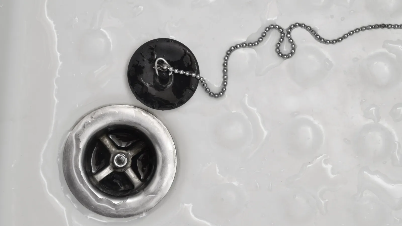 Close-up of a bathtub drain with a black rubber plug attached to a metal ball chain in a white tub.