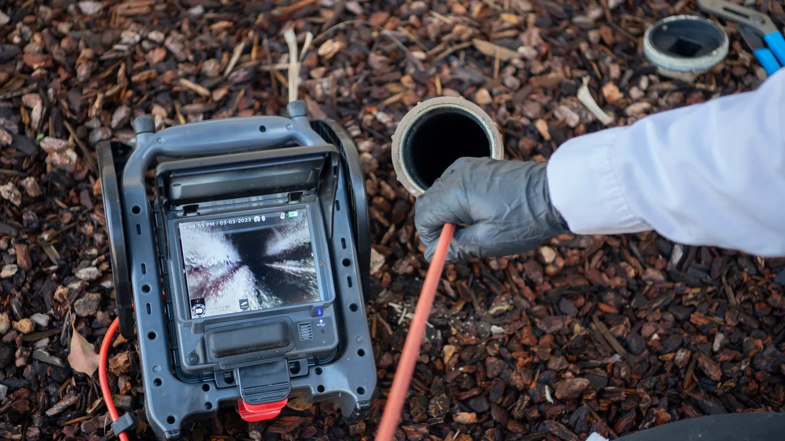 Technician using a sewer inspection camera to view inside a pipe surrounded by mulch and tools.